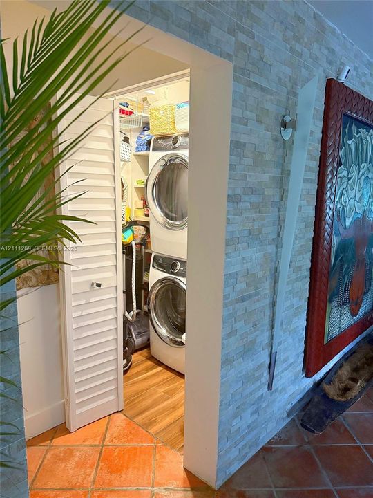 Laundry closet featuring white louvered doors, built-in shelving, and wood-finish flooring, contrasted by an exterior wall finished in stacked stone veneer