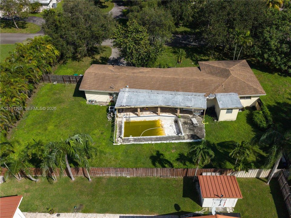 Aerial perspective of a single-story residence featuring a shingled roof, a covered concrete patio, an inground swimming pool, and a fenced backyard with mature landscaping