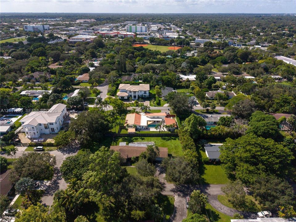 Aerial perspective featuring a residential lot with mature trees, established landscaping, and paved driveway access within a suburban setting