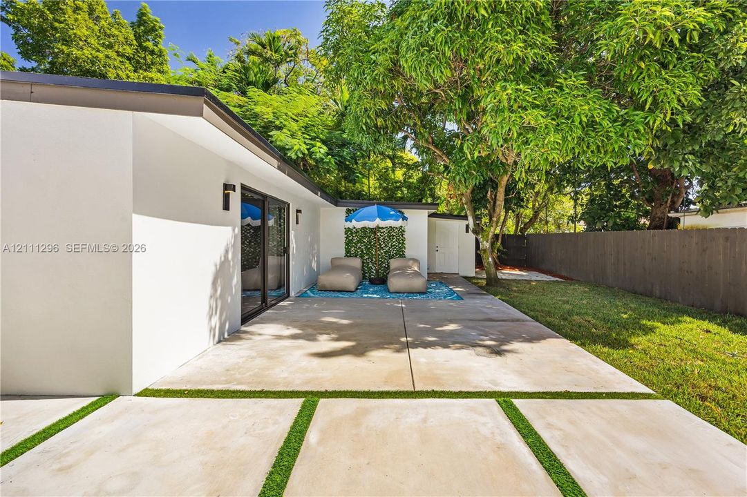 Concrete patio featuring inlaid synthetic turf strips, sliding glass doors, exterior wall-mounted lighting, and a fenced backyard with mature trees