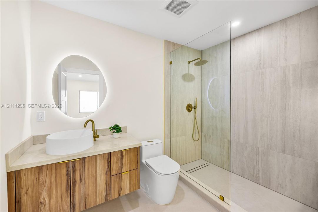 Floating wood-finish vanity with an oval vessel sink, back-lit circular mirror, walk-in shower with floor-to-ceiling tile, and brass-finish fixtures