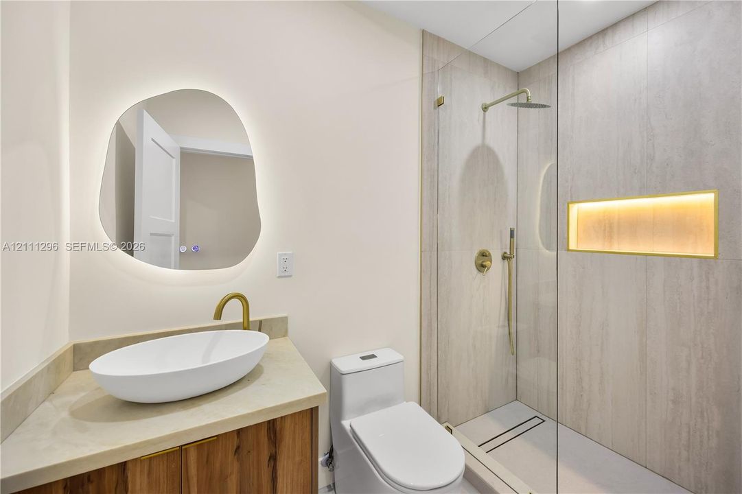 Bathroom featuring an asymmetrical backlit mirror, wood-finish vanity with a vessel sink, brass fixtures, glass-enclosed shower with tiled walls, and a recessed shower niche with integrated lighting