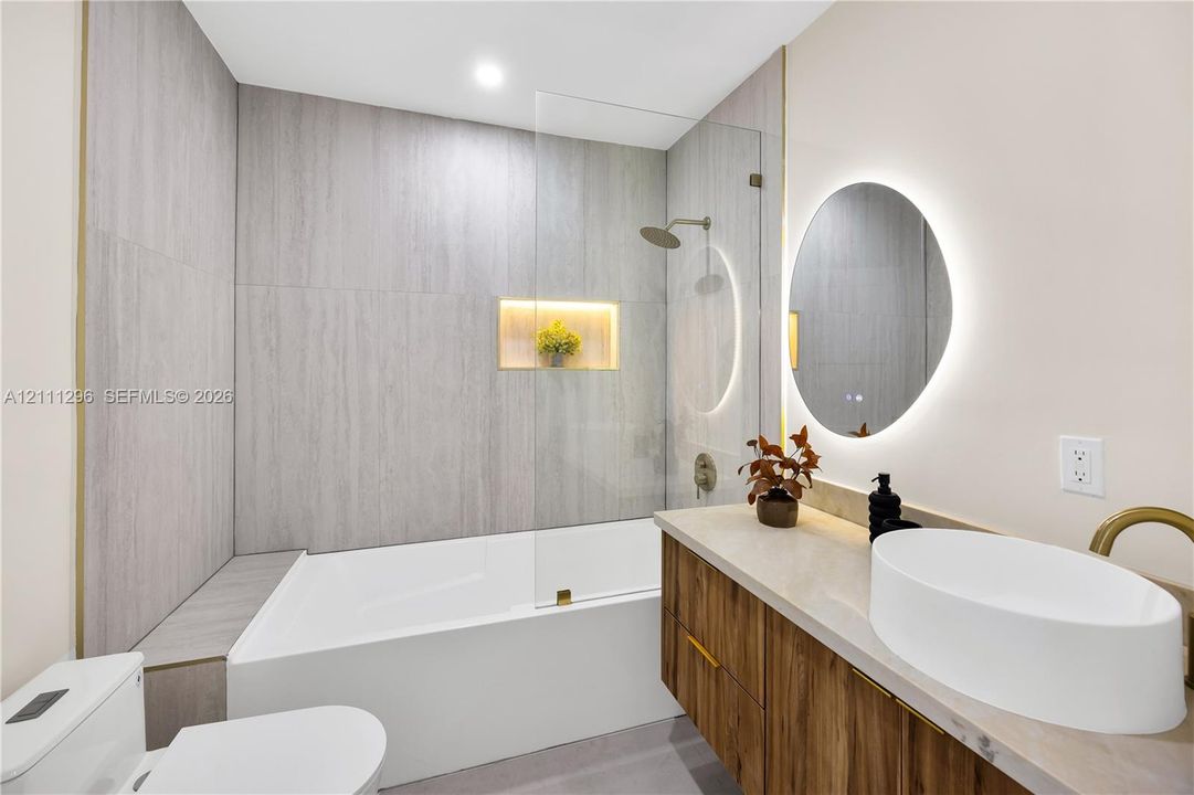 Bathroom featuring a wood-finish vanity with a vessel sink, quartz-style countertop, a walk-in shower with a glass panel and integrated recessed niche, and a circular LED-backlit mirror