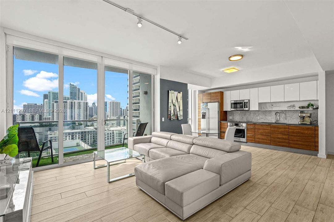 Open-concept living space with wood-finish flooring, floor-to-ceiling glass doors, built-in dual-tone cabinetry, stainless steel appliances, and integrated lighting