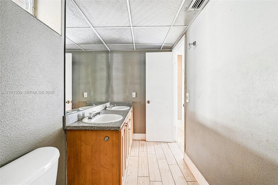 Dual-sink vanity with a solid surface countertop, wood-finish cabinetry, full-width wall mirror, and neutral tile flooring