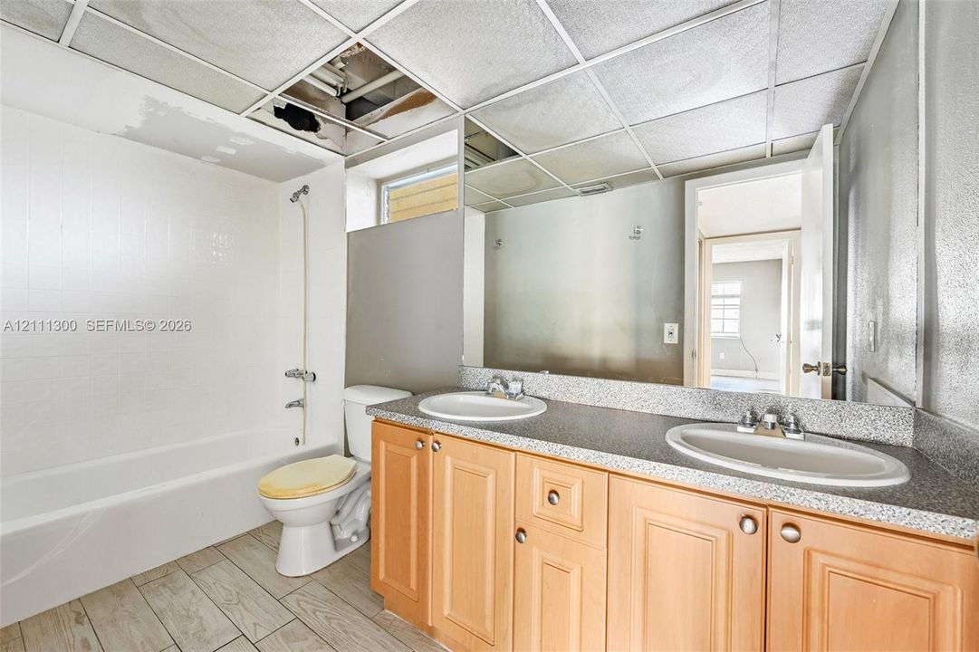 Dual-sink vanity with light-tone wood cabinetry, solid-surface countertop, full-width wall mirror, and tile-finish flooring