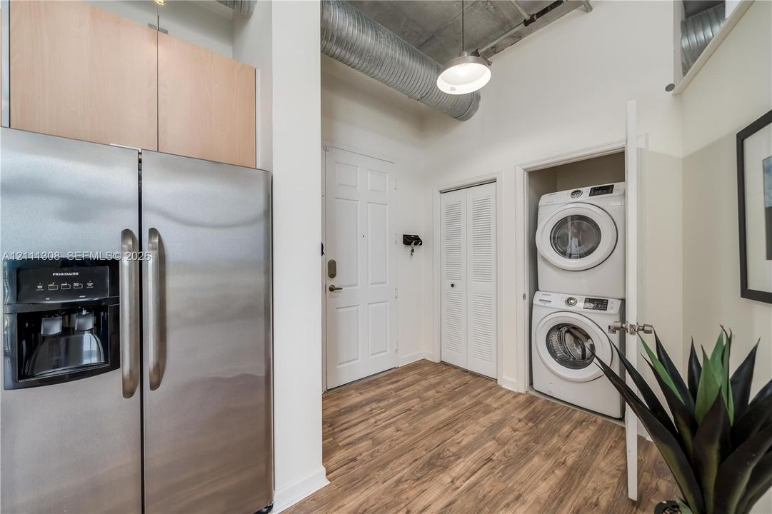 Stainless steel refrigerator, wood-finish flooring, exposed industrial ductwork, high ceilings, and stacked laundry unit