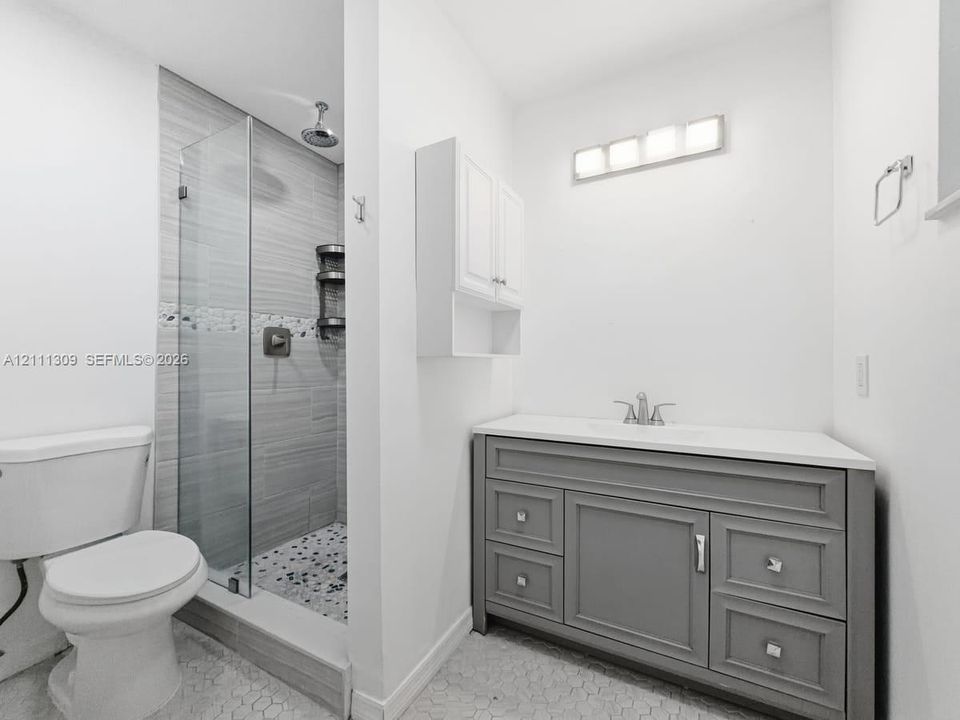 Bathroom featuring a grey vanity with a solid surface countertop, glass-enclosed walk-in shower with horizontal tile walls, pebble-style floor tiling, and wall-mounted cabinetry