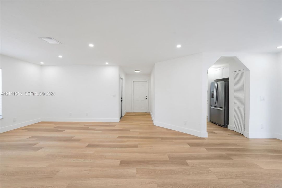 Open-concept interior featuring light wood-finish flooring, recessed lighting, smooth white walls, and a stainless steel refrigerator