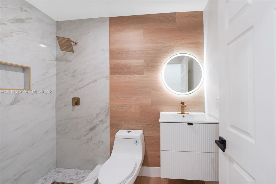 Bathroom featuring a walk-in shower with large-format tile surround, wood-finish wall paneling, a floating vanity with a fluted cabinet front, a backlit circular mirror, and brass-tone fixtures