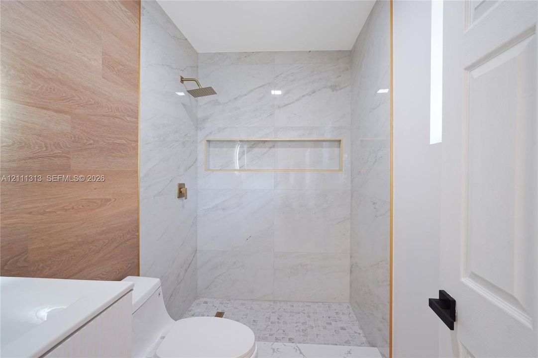 Walk-in shower featuring large-format light-veined tile, wood-finish accent wall, built-in recessed shelf, rainfall shower head, and decorative metallic trim