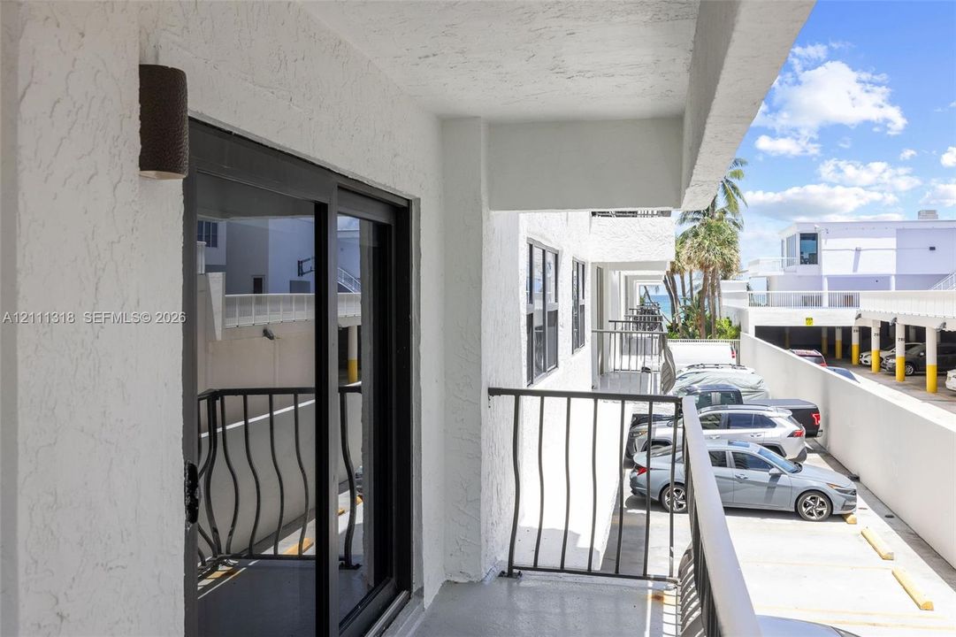 Covered exterior balcony with textured stucco walls, black metal railing, sliding glass door, and cylindrical wall-mounted exterior light fixture