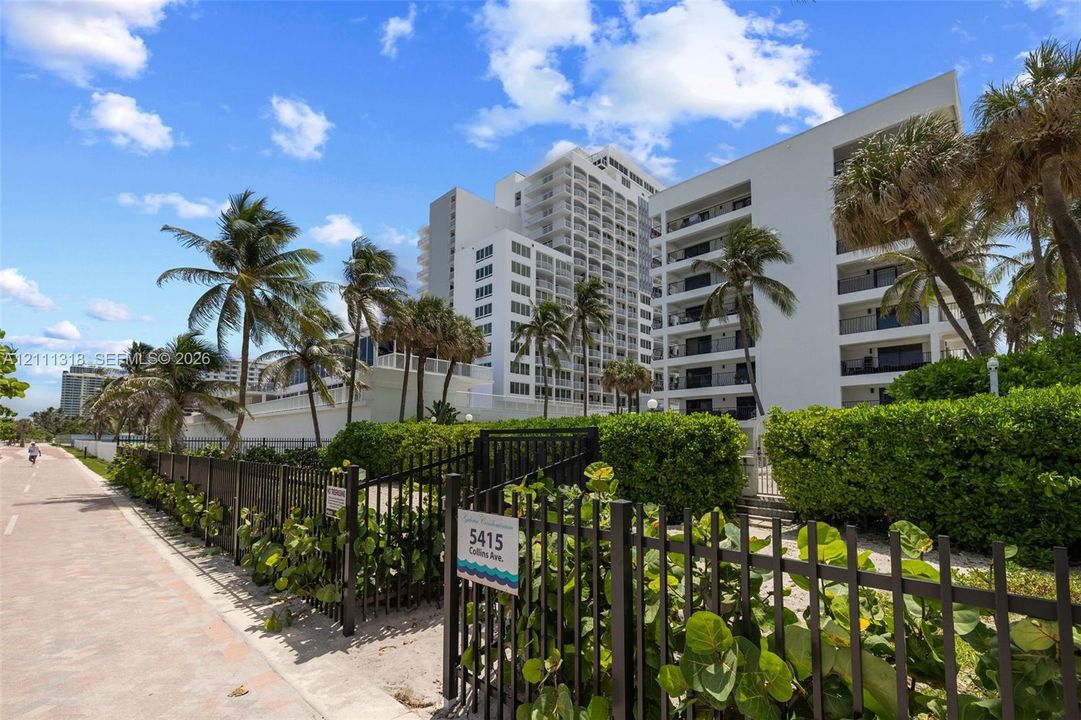 Multi-story building featuring white stucco exterior, stacked cantilevered balconies, metal perimeter fencing, and established tropical landscaping