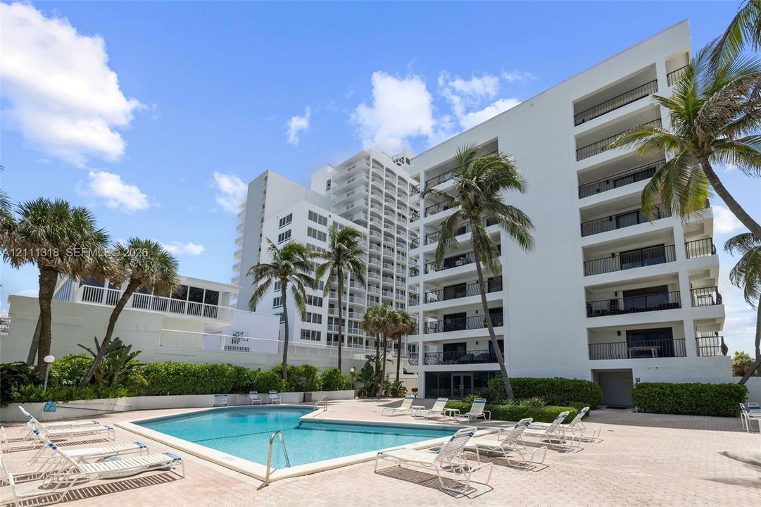 Multi-story residential complex featuring private balconies, an inground swimming pool, paved sundeck, and mature palm trees