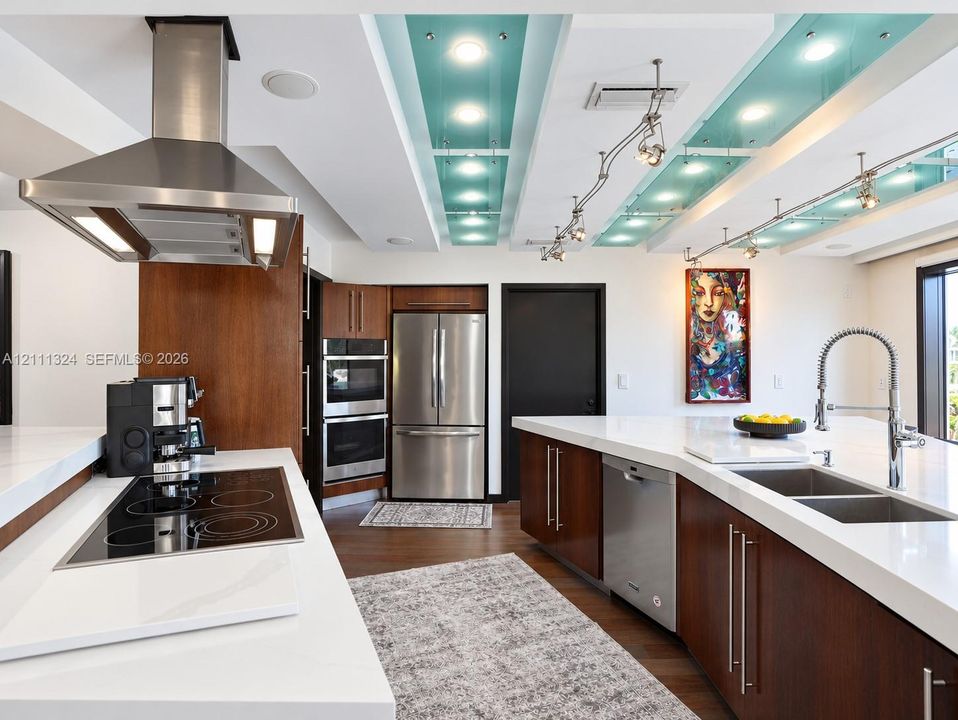 Kitchen featuring wood-veneer cabinetry, quartz countertops, a stainless steel chimney hood, built-in double ovens, and a recessed ceiling installation with decorative glass panels