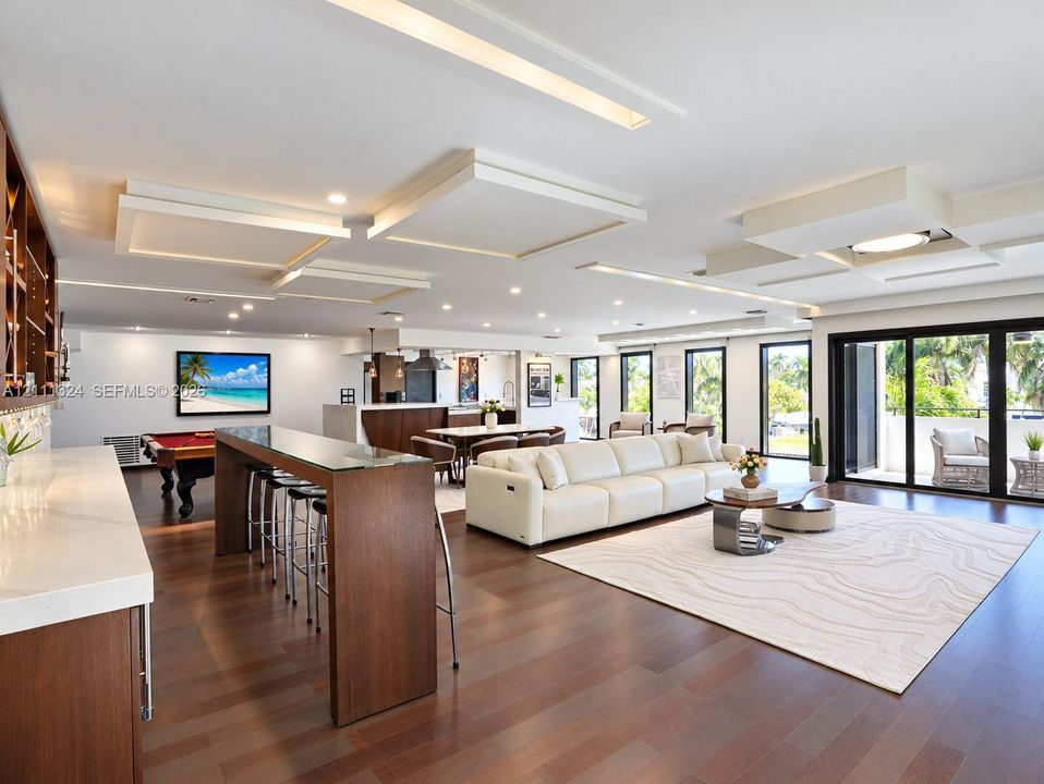 Open-concept living space featuring wood-finish flooring, custom coffered ceiling with recessed lighting, built-in bar with stone countertop, and floor-to-ceiling sliding glass doors