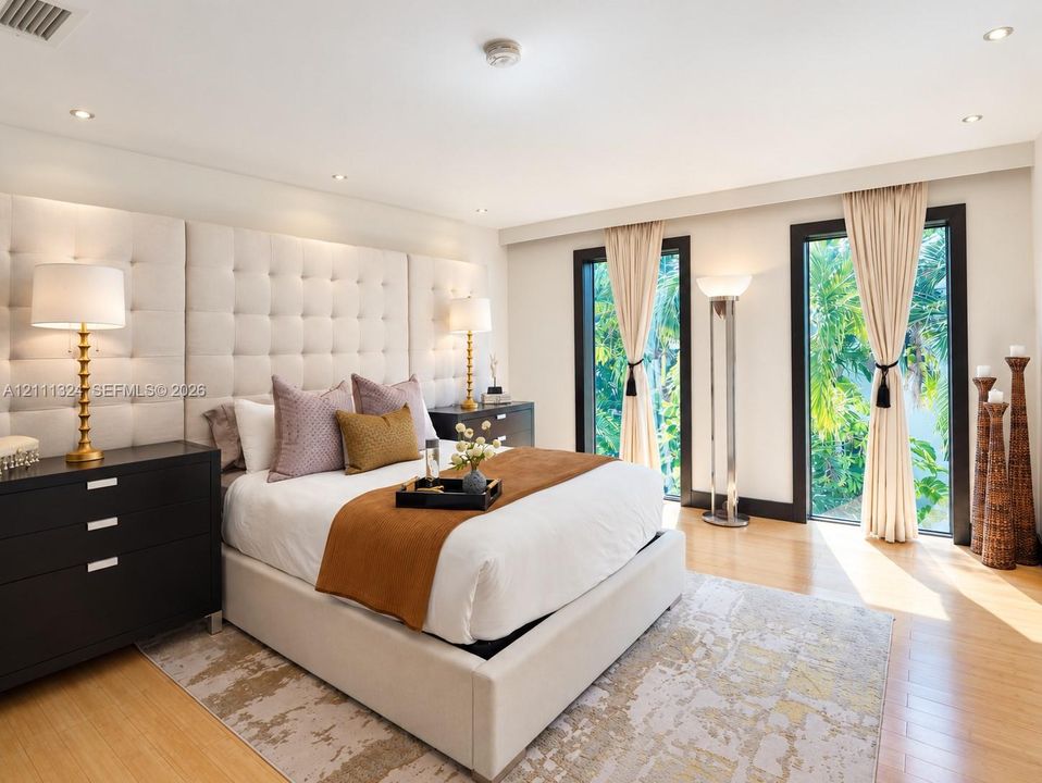 Floor-to-ceiling tufted fabric headboard, recessed ceiling lighting, and two full-length glass floor to ceiling windows with dark frames and integrated drapery