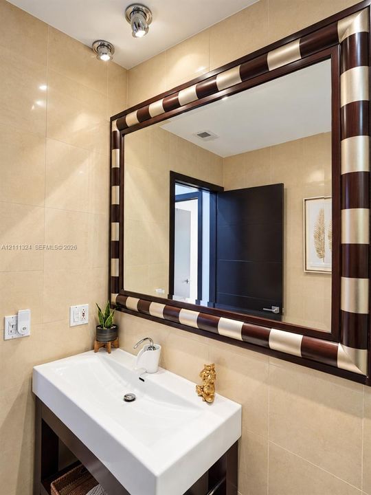 Rectangular white vessel sink atop a  wood-finish vanity, large wall mirror with a decorative two-tone wood and metal frame, and full-height stone wall tile