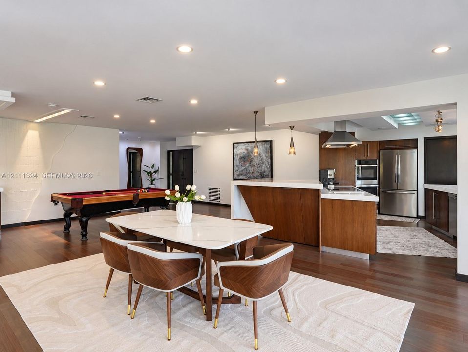 Open-concept kitchen featuring flat-panel wood cabinetry, integrated stainless steel appliances, a stainless steel range hood, and a kitchen island with a stone countertop, all illuminated by recessed lighting