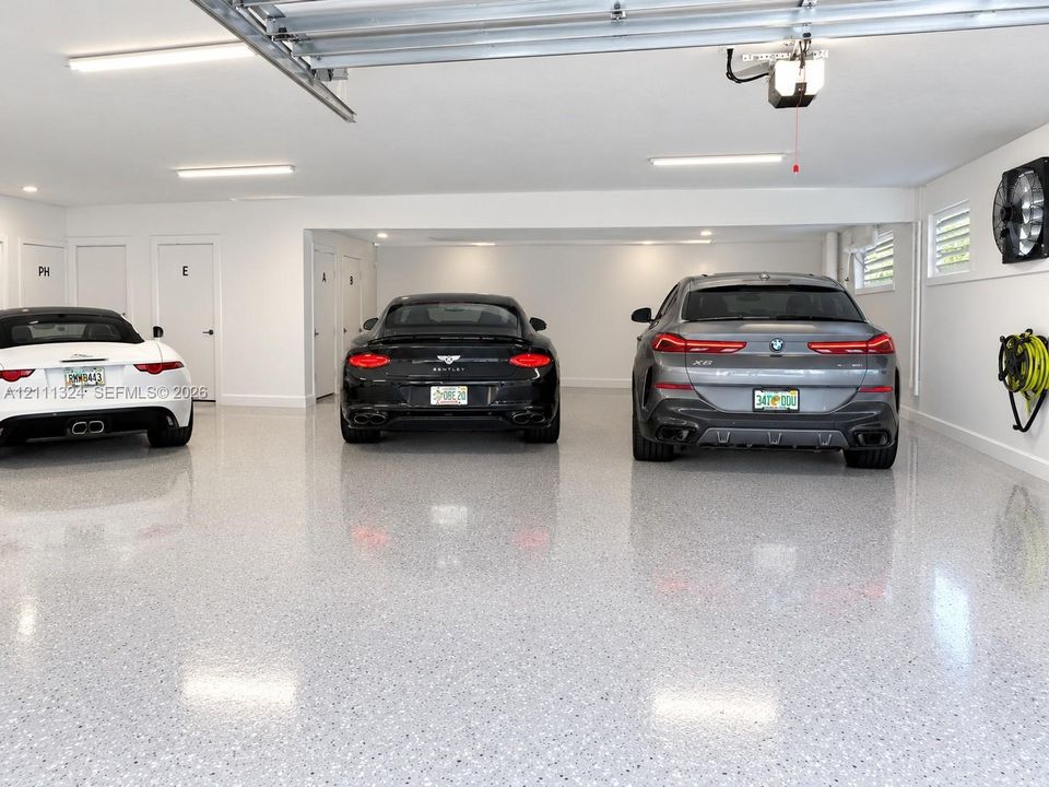 Spacious three-car garage featuring finished epoxy flooring, white walls, overhead fluorescent lighting, and multiple interior access doors