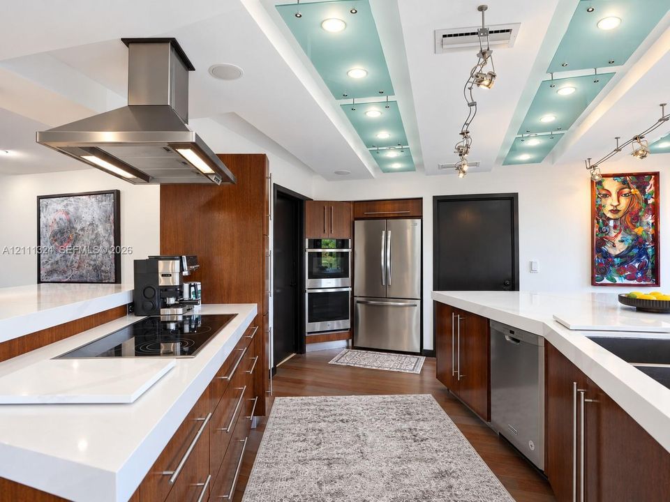 Kitchen featuring custom wood cabinetry, stainless steel appliances including a built-in double oven and range hood, white solid-surface countertops, and recessed lighting with decorative glass panels