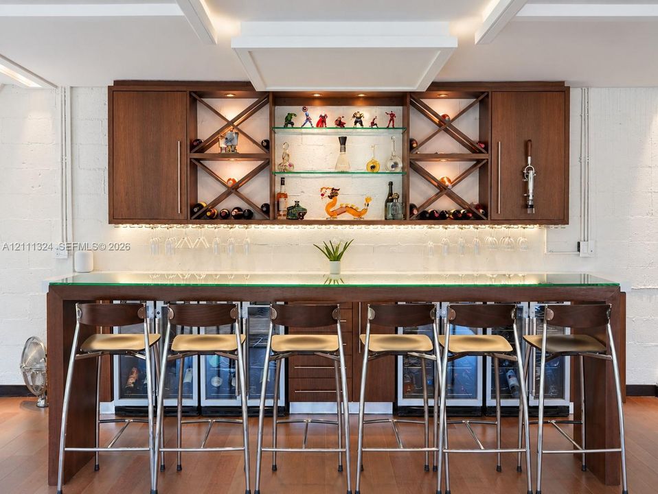 Custom built-in bar featuring wood cabinetry, integrated glass shelving with X-frame detailing, under-cabinet lighting, and a glass-top counter with multiple under-counter refrigeration units