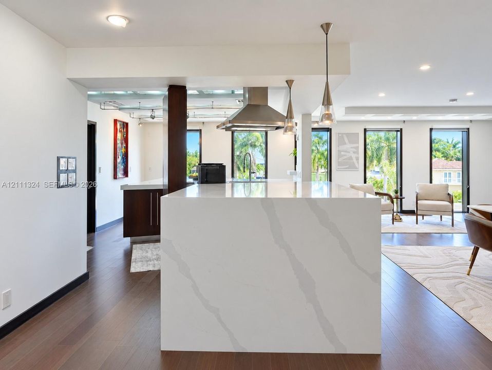 Waterfall kitchen island with stone countertop, wood-finish flooring, stainless steel range hood, pendant lighting, and dark-toned cabinetry
