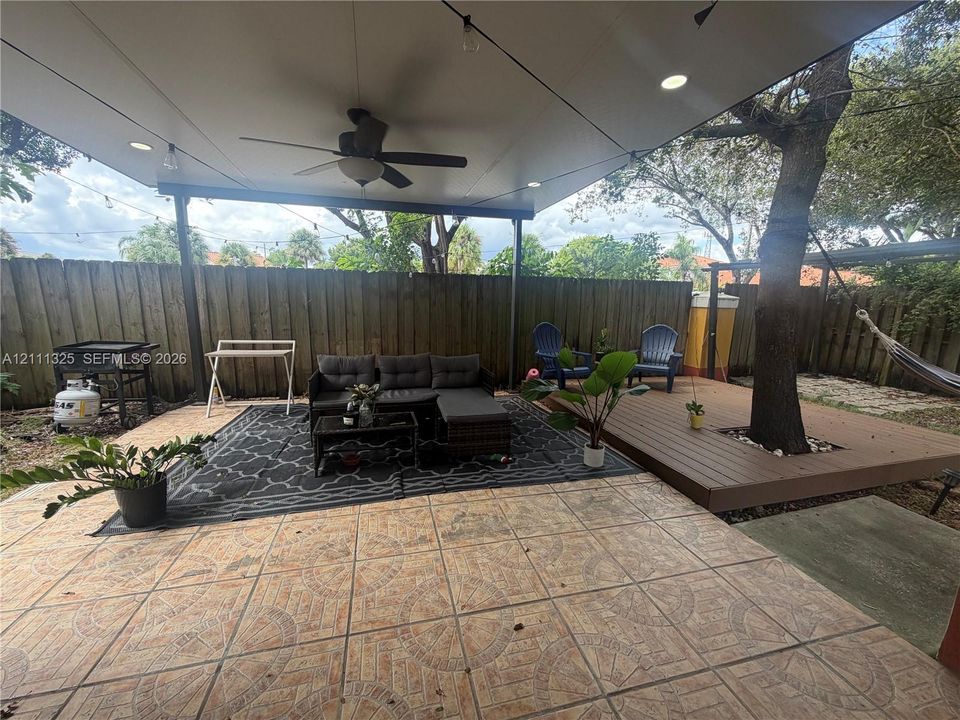 Covered patio featuring patterned tile flooring, recessed lighting, and a ceiling fan, complemented by an elevated wood-finish deck surrounding a mature tree