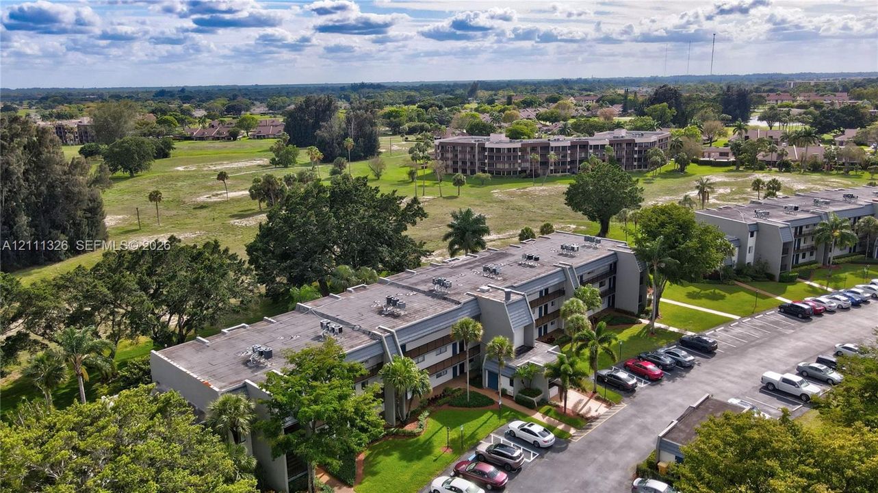 Aerial perspective featuring multi-story residential buildings, paved surface parking, manicured landscaping, and mature trees.