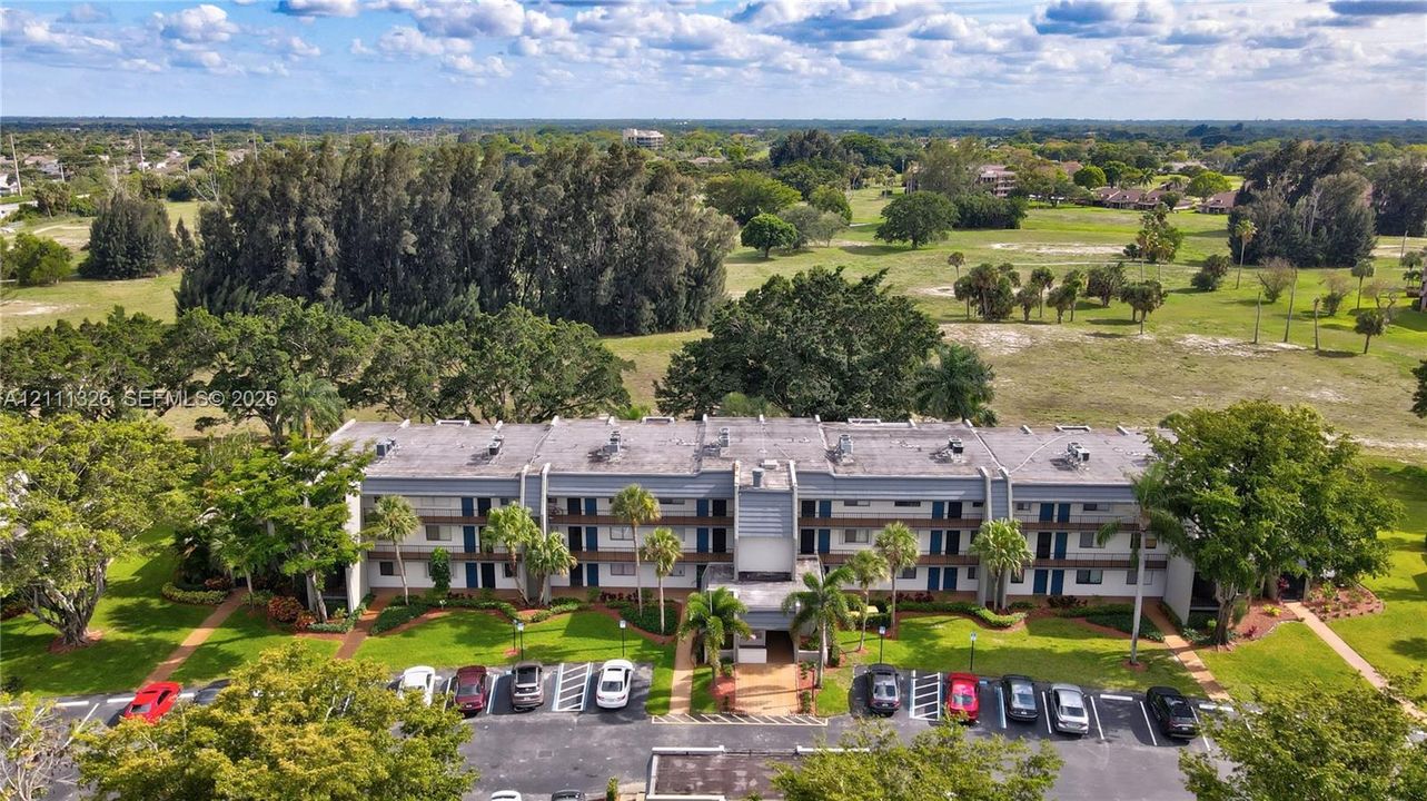 Multi-story residential building featuring a concrete facade, integrated parking area, and surrounding green space with mature trees.