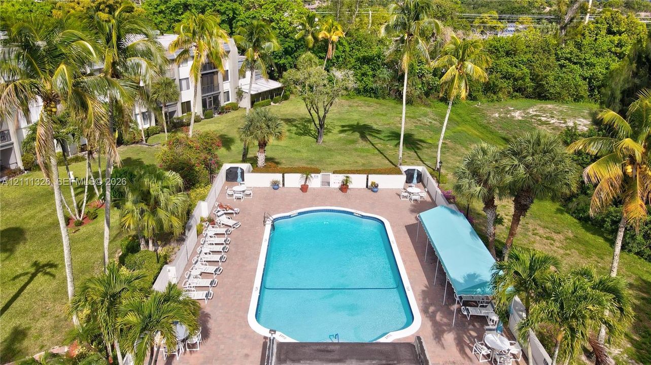 Outdoor community swimming pool featuring a paver deck, perimeter fencing, and a covered patio area set within a landscaped property with mature trees.