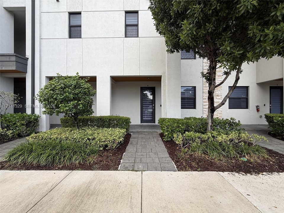 Modern facade with panelized exterior, multi-pane entry door, stone-clad accent wall, stone paver walkway, and manicured shrubbery