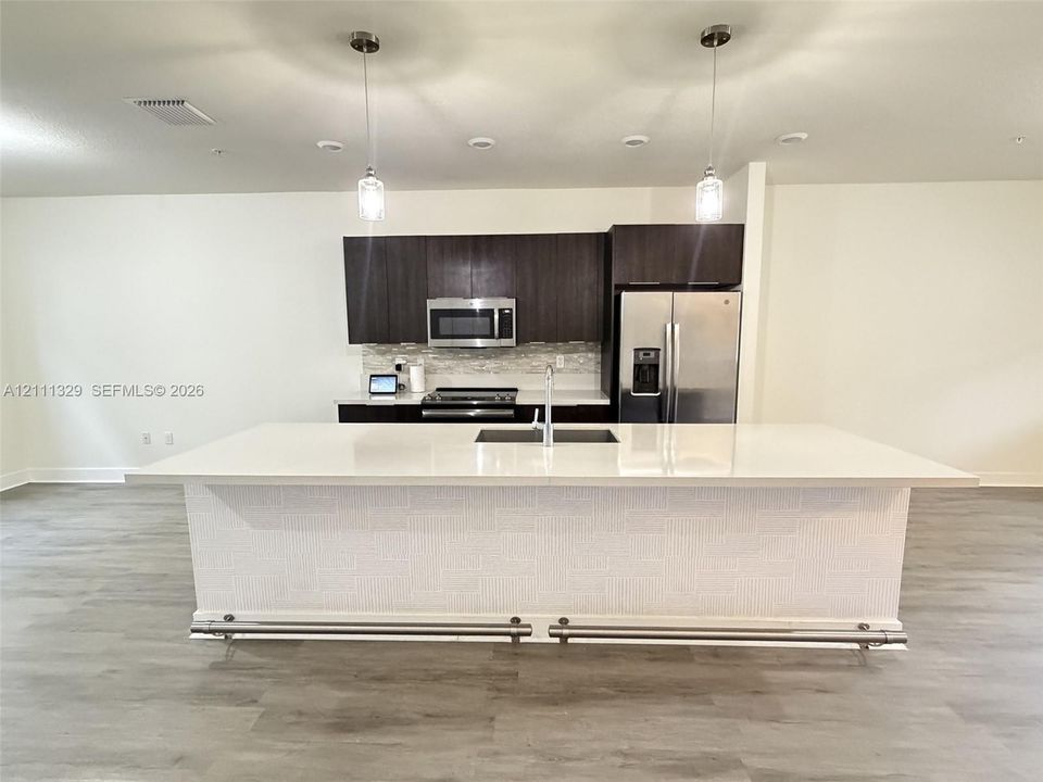 Kitchen featuring an expansive island with a solid-surface countertop, dark cabinetry, stainless steel appliances, textured island base finish, and pendant lighting