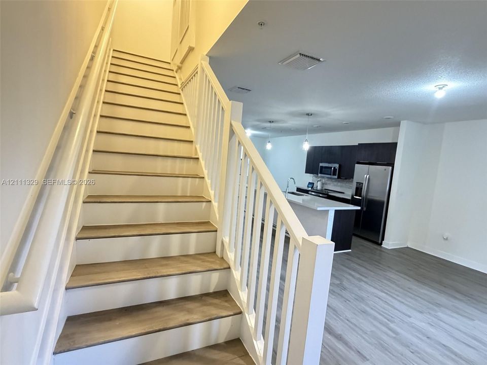 Staircase with wood-finish treads and white balusters, leading to an open-concept kitchen featuring dark cabinetry, an island with a light-toned countertop, and stainless steel appliances