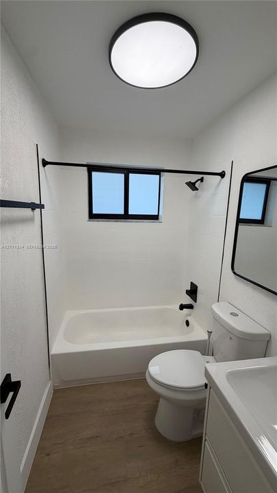 Bathroom featuring wood-finish flooring, a white vanity with an integrated countertop sink, matte black hardware, a shower-tub combination with white wall surround, and a circular flush-mount ceiling light