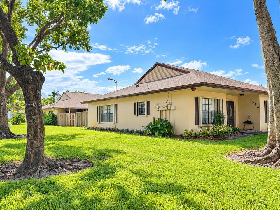 Single-story residence featuring light-toned stucco exterior, shingled hip roof, manicured lawn, and mature trees