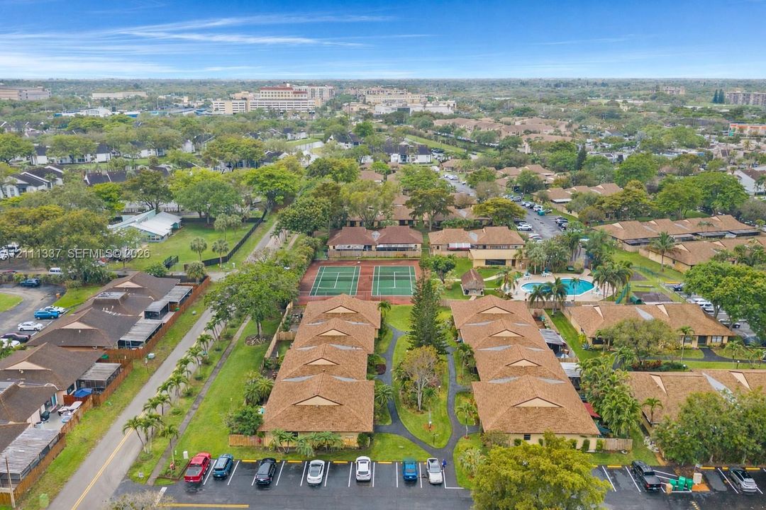 Aerial perspective of a multi-unit residential community featuring brown shingled roofing, dual tennis courts, a swimming pool, and dedicated paved parking areas