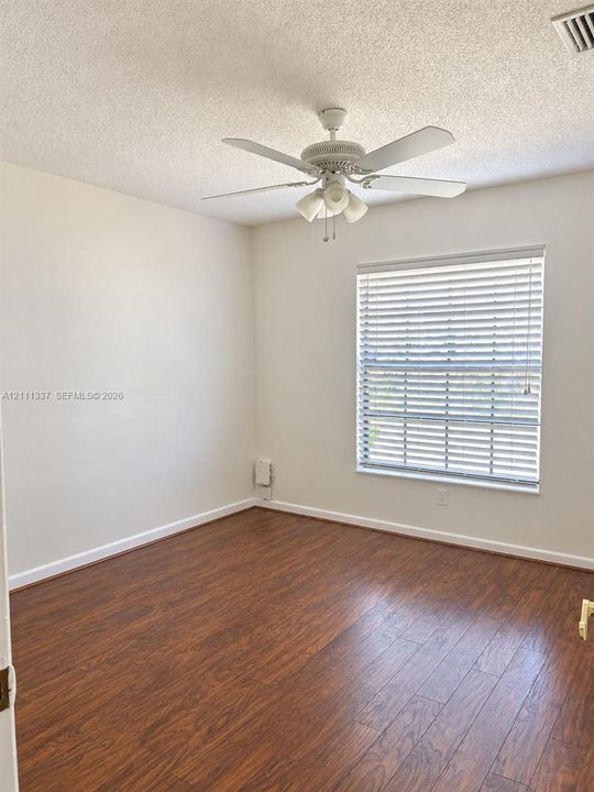 Wood-finish flooring, neutral wall tones, multi-blade ceiling fan with integrated light fixture, and horizontal window blinds