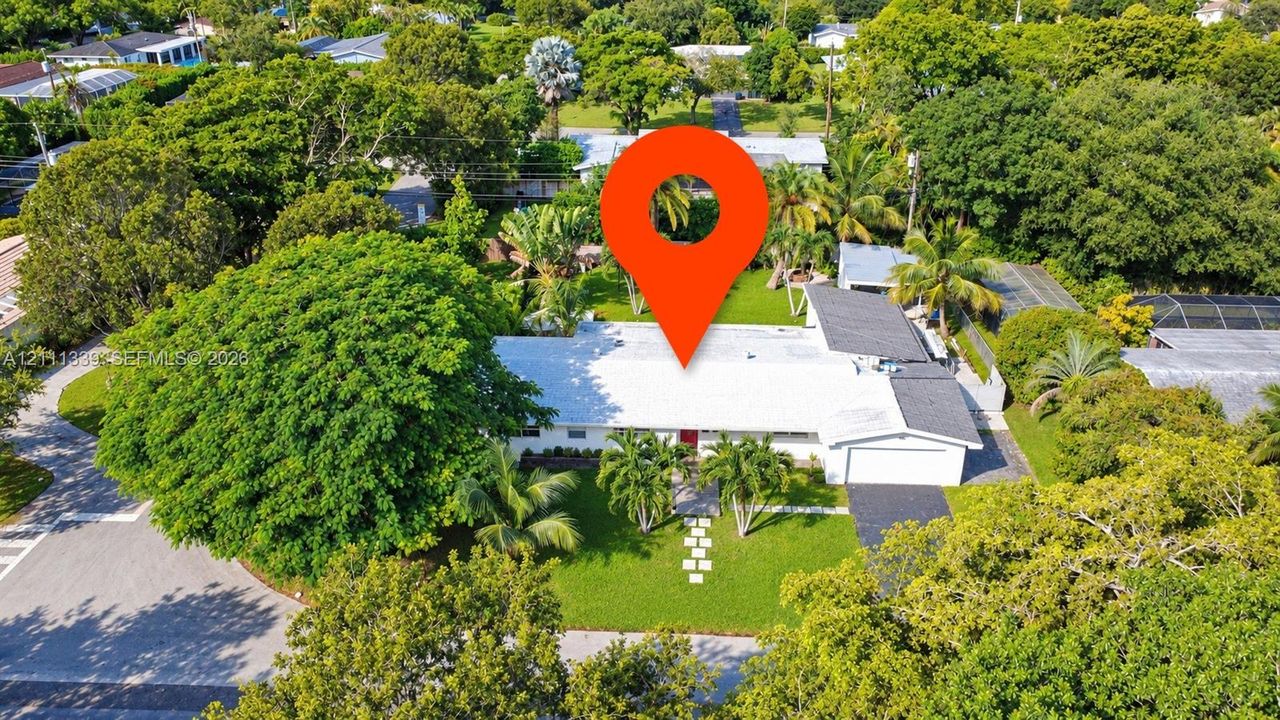 Aerial perspective of a single-story residence featuring a shingled roof, an attached garage, a paved driveway, and established landscaping with mature trees on a spacious lot