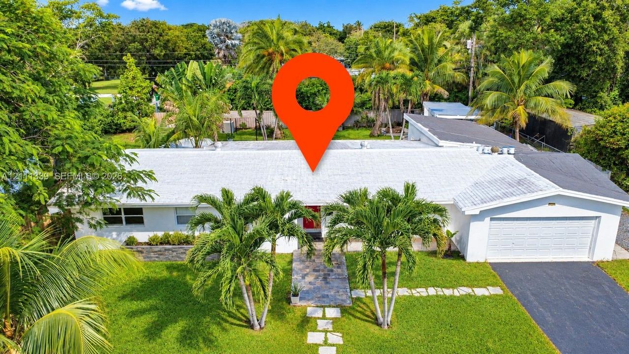 Single-story residence featuring a white exterior, attached two-car garage, asphalt driveway, stone paver walkway, and manicured landscaping with mature palm trees