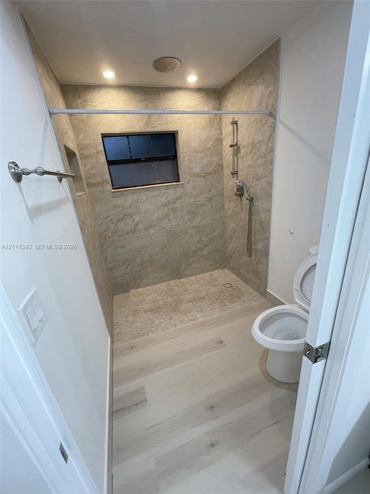 Walk-in shower featuring floor-to-ceiling stone-finish tile surround, recessed wall niche, adjustable handheld shower fixture, and light-toned wood-finish flooring