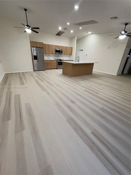 Open-concept area featuring light-toned wood-finish flooring, vaulted ceilings with recessed lighting, a central island with a stone-surface countertop, stainless steel appliances, and shaker-style cabinetry