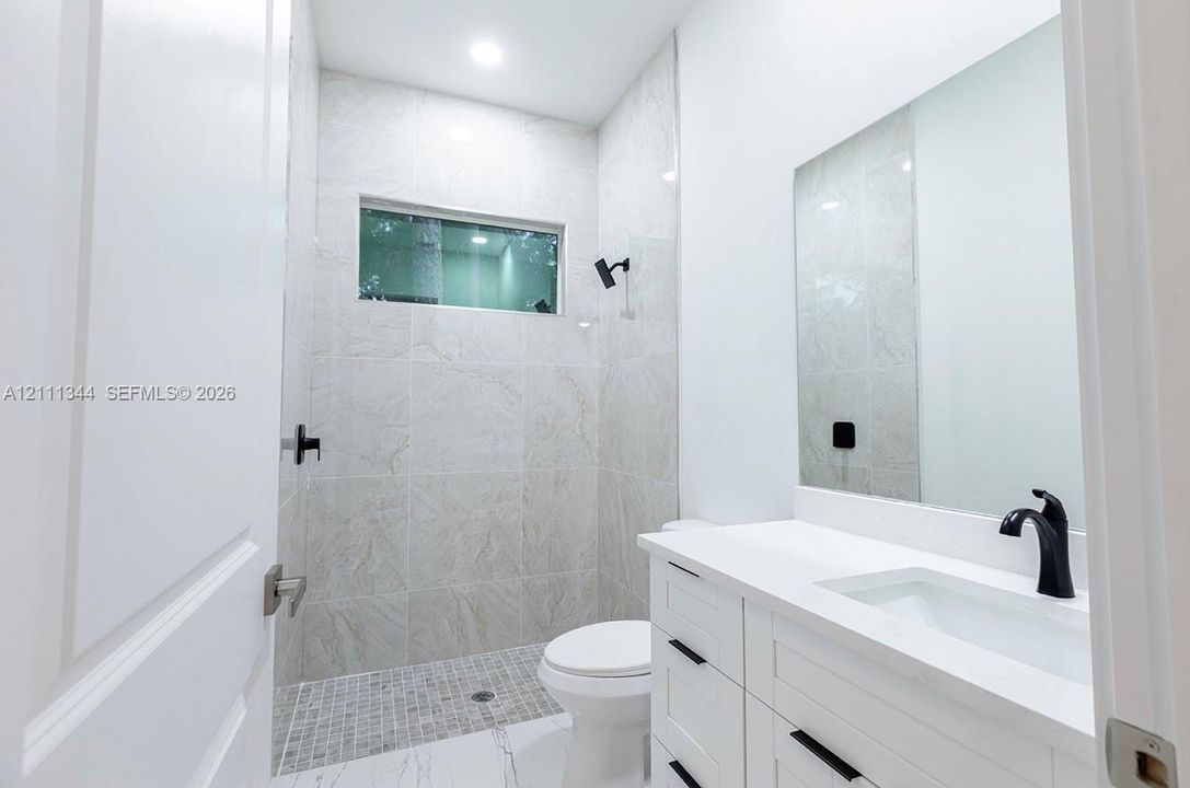 Bathroom featuring a walk-in shower with stone-finish tile surround, a white vanity with a composite countertop, an undermount sink with matte black hardware, and an expansive wall-mounted mirror