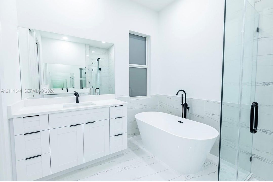 Freestanding soaking tub with matte black floor-mounted faucet, white cabinetry with quartz countertop, integrated rectangular sink, large wall-mounted mirror, and glass-enclosed shower with tiled surround