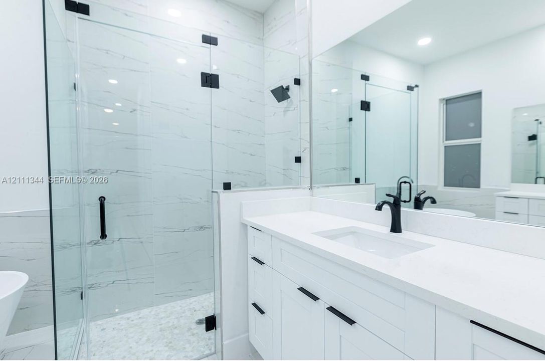 Walk-in shower with floor-to-ceiling tile, frameless glass enclosure, and matte black fixtures, paired with a white vanity featuring a stone-surface countertop and matte black hardware