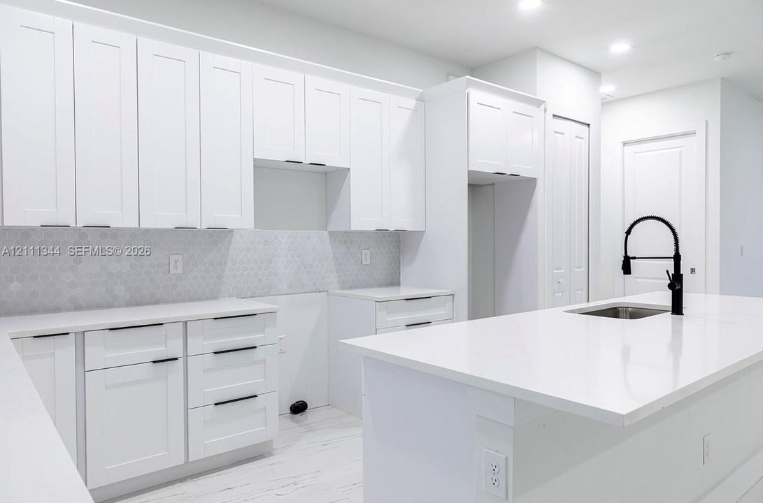 Kitchen featuring white shaker-style cabinetry, a central island with an undermount sink, a matte black gooseneck faucet, light-toned stone-finish countertops, and a hexagonal tile backsplash
