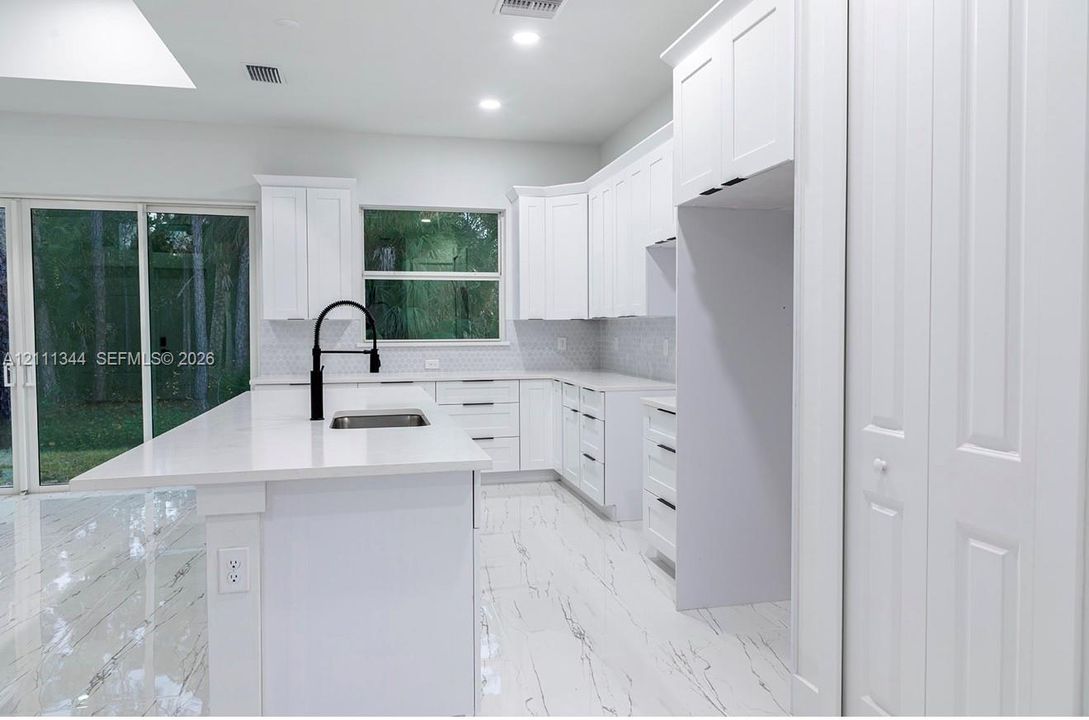 Kitchen featuring white shaker-style cabinetry, a quartz-topped island with an undermount stainless steel sink, matte black hardware, stone-patterned tile flooring, and a glass-paneled sliding door