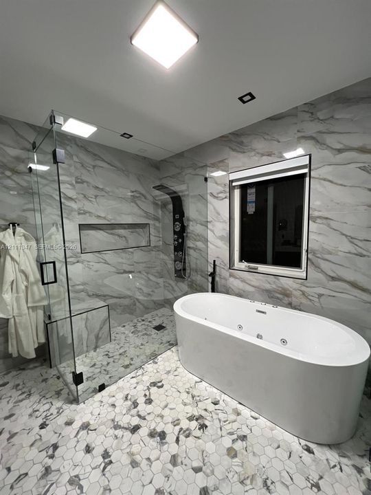 Freestanding jetted tub, glass-enclosed walk-in shower with integrated bench and wall-mounted shower panel, floor-to-ceiling stone-finish wall tile, and hexagonal tile flooring
