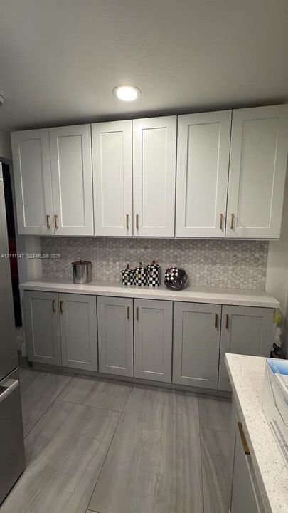 Kitchen cabinetry featuring light-toned Shaker-style doors, stone-composite countertops, hexagonal tile backsplash, metallic hardware, and large-format tile flooring