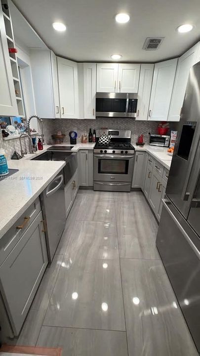 Kitchen featuring white cabinetry, light-toned stone-finish countertops, stainless steel appliances, large-format tile flooring, and a farmhouse-style sink